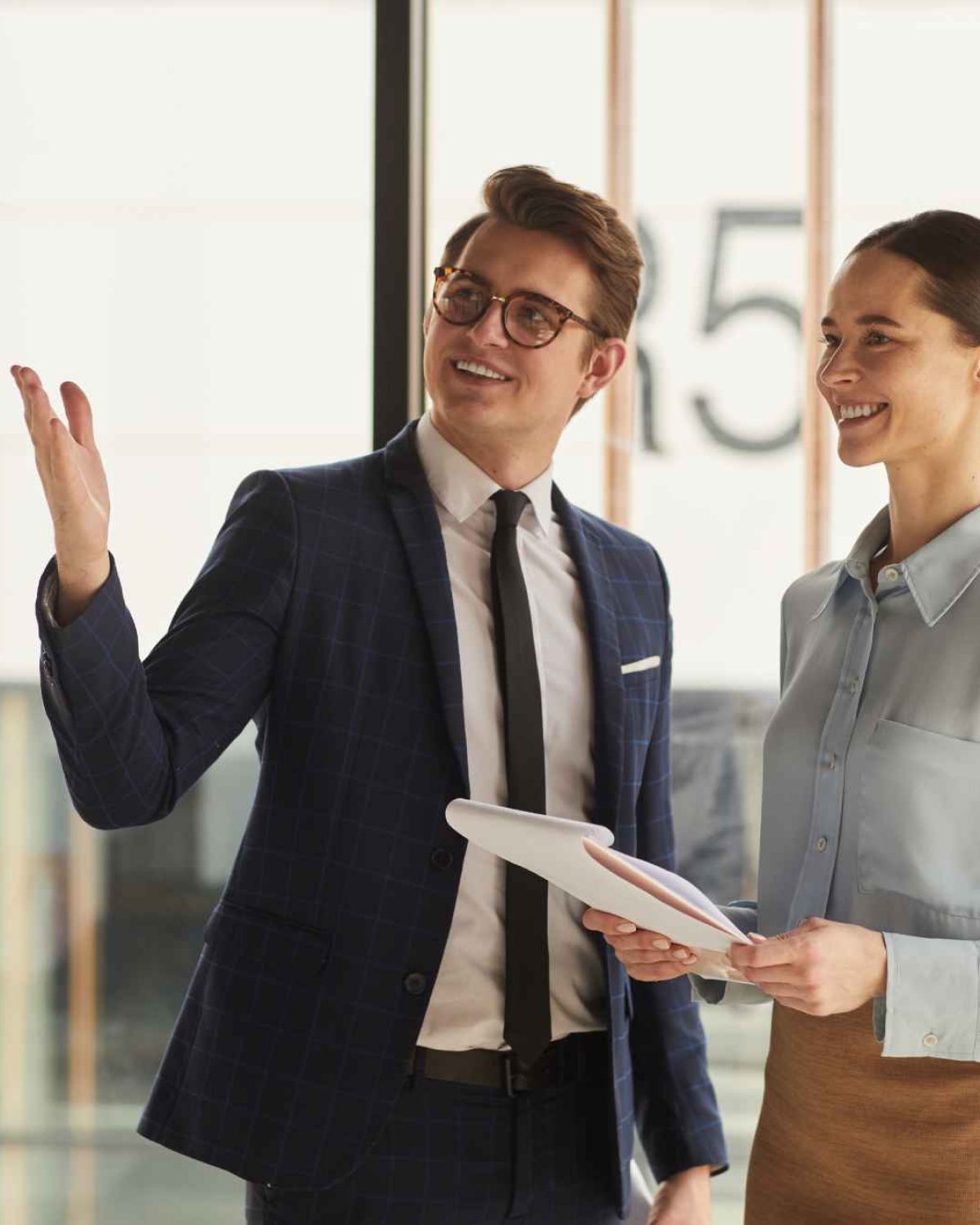 Waist up portrait of smiling real estate agent discussing property with female client and pointing up while standing in empty office building interior lit by sunlight, copy space